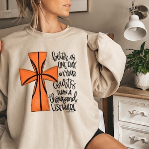 May include: A beige sweatshirt with a basketball-shaped cross graphic and the text "better is one day in your courts than a thousand elsewhere Psalm 84:10".