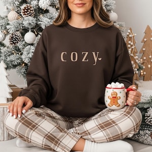May include: A dark brown sweatshirt with the word "COZY" printed in a light brown font. The person is wearing plaid pants and holding a mug with a gingerbread man design. The mug is filled with marshmallows.
