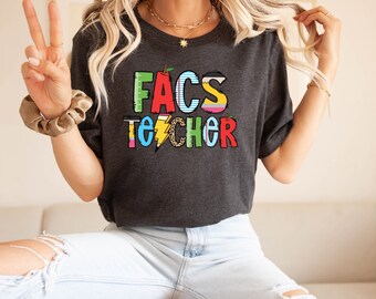 May include: A dark gray t-shirt with the text "FACS TEACHER" in colorful letters. The letters are decorated with various school-related graphics, including an apple, a pencil, a ruler, and a lightning bolt.