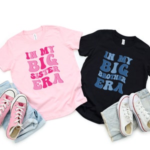 Può includere: Due t-shirt con il testo "In My Big Sister Era" e "In My Big Brother Era" in un font retrò. Le magliette sono rosa e nere con testo blu e rosa.