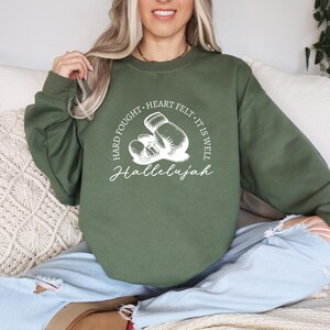 May include: Olive green sweatshirt with a white graphic of boxing gloves and the words "Hard Fought, Heart Felt, It Is Well, Hallelujah". The sweatshirt is a crew neck style.
