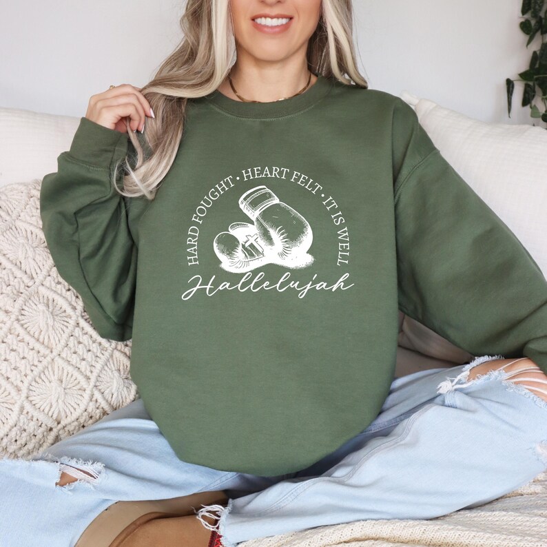 May include: Olive green sweatshirt with a white graphic of boxing gloves and the words "Hard Fought, Heart Felt, It Is Well, Hallelujah". The sweatshirt is a crew neck style.