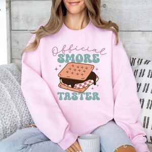 May include: A pink sweatshirt with the text "Official S'more Taster" in a retro font. A cartoon illustration of a s'more with chocolate and marshmallow is above the text.