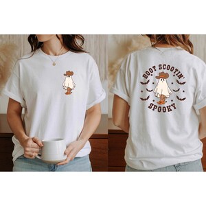 May include: White t-shirt featuring a cartoon ghost wearing a cowboy hat and boots. The front design shows the ghost, while the back displays the text "Boot Scootin' Spooky" with bats and stars. A fun, Halloween-themed graphic tee.