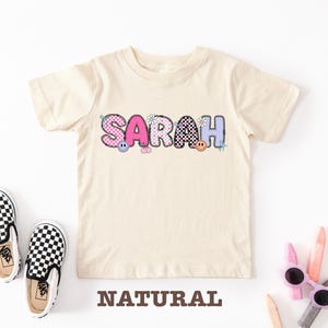 Può includere: Una maglietta color crema con il nome "SARAH" in lettere colorate e decorate. Le lettere presentano motivi rosa, blu e a scacchi neri, con faccine sorridenti e fiori. Scarpe slip-on a scacchi bianchi e neri sono accanto alla maglietta. La parola "NATURAL" è stampata sotto.