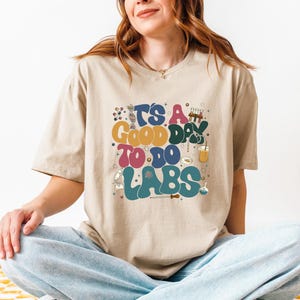 May include: A beige t-shirt with a colorful graphic that says "It's a good day to do labs". The graphic features a beaker, test tubes, a microscope, and other science-related imagery.