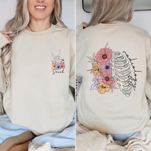 Pode incluir: Uma camisa de moletom bege com um design floral e uma caixa tor&aacute;cica. O texto "radiologia" est&aacute; escrito verticalmente do lado direito do design. O texto "Sarah" est&aacute; escrito na frente da camisa de moletom.