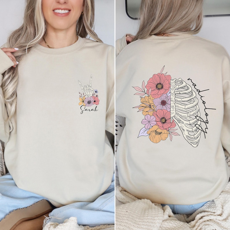 Pode incluir: Uma camisa de moletom bege com um design floral e uma caixa tor&aacute;cica. O texto "radiologia" est&aacute; escrito verticalmente do lado direito do design. O texto "Sarah" est&aacute; escrito na frente da camisa de moletom.