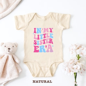 Può includere: Un body per neonato beige a maniche corte con chiusura a pressione. Il body ha una grafica rosa, blu e bianca che dice "In My Little Sister Era".