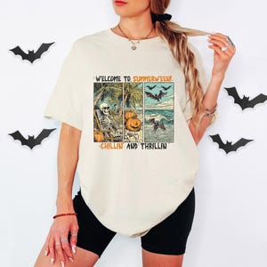 May include: Cream-colored t-shirt with a graphic that reads "WELCOME TO SUMMERWEEN!" and "CHILLIN AND THRILLIN". The graphic features a skeleton, pumpkins, bats, and a beach scene. Black shorts are also visible.