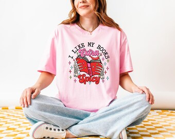 May include: A light pink t-shirt with the text "I Like My Books Extra Spicy" and a graphic of a book held by skeleton hands. The book has a red cover and a chili pepper. The person is wearing light blue jeans and white sneakers.