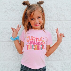 May include: A pink t-shirt with a colorful graphic that says "Little Miss Three-nager".