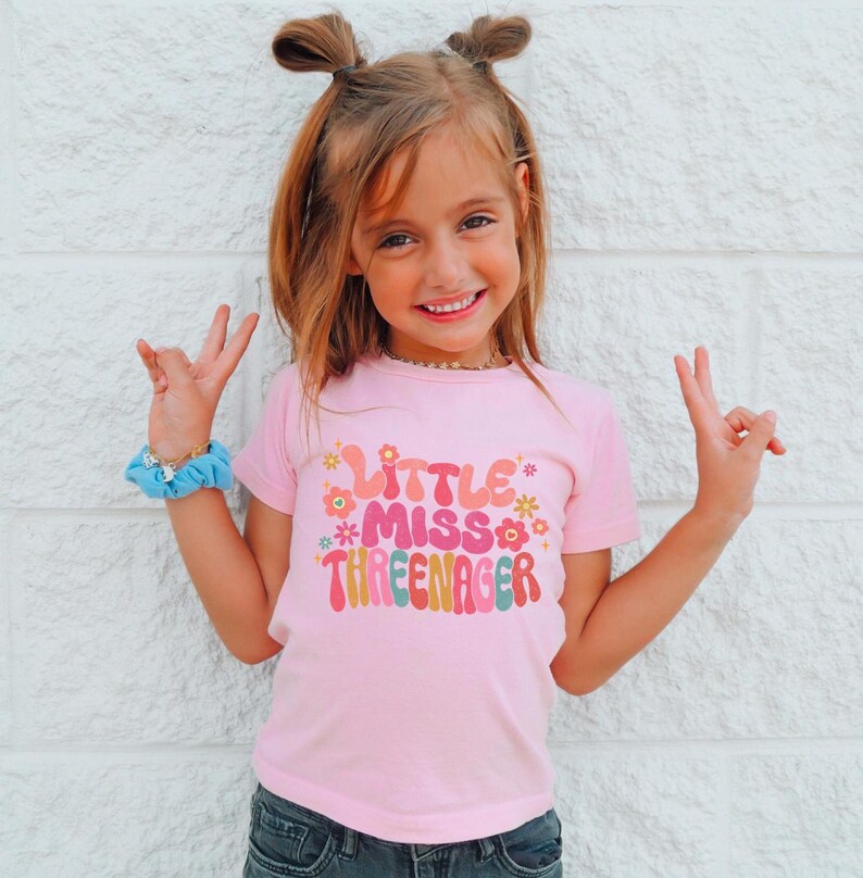 May include: A pink t-shirt with a colorful graphic that says "Little Miss Three-nager".