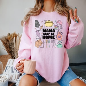 May include: Pink sweatshirt with a graphic design of kitchen utensils and the text "Mama Stay at Home".