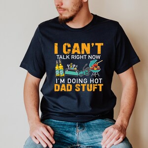 May include: A navy blue t-shirt with the text "I can't talk right now I'm doing hot dad stuff" in orange. The shirt features a graphic of two bottles of beer, a lawnmower, tools, and a grill with hot dogs and hamburgers.