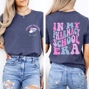 May include: Navy blue t-shirt with the text "FUTURE PHARMD" and a pill graphic on the front. The back of the shirt features the phrase "IN MY PHARMACY SCHOOL ERA" in pink and blue lettering. The shirt is paired with blue jeans.