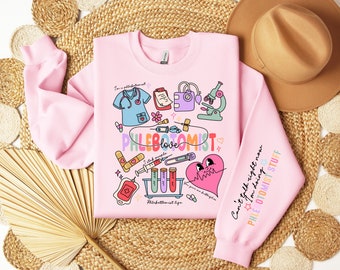 May include: A pale pink sweatshirt featuring phlebotomy-themed illustrations. The design includes a medical scrub top, microscope, and a heart. Text on the sweatshirt reads "PHLEBOTOMIST love" and "Can't talk right now I'm doing PHLEBOTOMIST stuff."