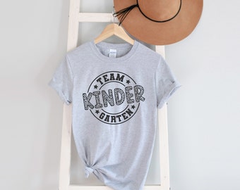 May include: Gray t-shirt with a black and white leopard print circle design that says "Team Kindergarten" in black text.