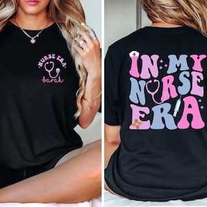May include: Black t-shirt with a pink and blue graphic that says "In My Nurse Era" with a stethoscope, bandage, and syringe. The front of the shirt has a pink stethoscope graphic with the text "Nurse Era" and a name.