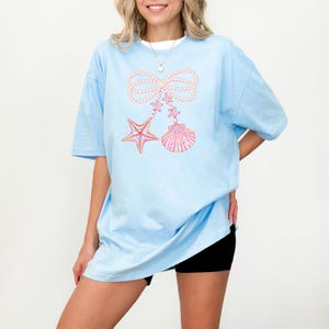 May include: A light blue oversized t-shirt with a graphic of a pearl bow, a starfish, and a seashell. The graphic is in pink, yellow, and blue.