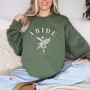 May include: A green sweatshirt with the word "ABIDE" in white lettering, a small branch with leaves and berries, and the text "JOHN 15" underneath.