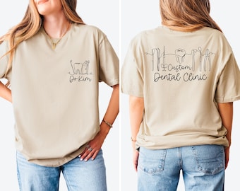 May include: Beige t-shirt with black line art of dental tools and a tooth. The front of the shirt has the text "Dr. Kim" below a tooth and dental tool design. The back of the shirt has the text "Custom Dental Clinic".