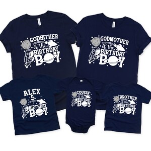 May include: Five navy blue t-shirts with white astronaut and planet designs. The shirts say "Godfather of the Birthday Boy", "Godmother of the Birthday Boy", "Alex 5 Birthday Boy", "Cousin of the Birthday Boy", and "Brother of the Birthday Boy".