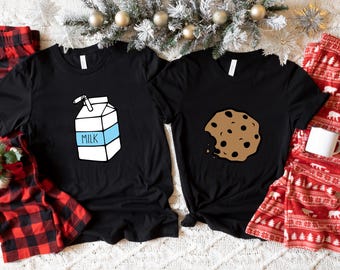May include: Two black short-sleeve t-shirts. One shirt features a white milk carton graphic with a blue band and the word MILK. The other shirt features a brown chocolate chip cookie graphic with a bite taken out of the side of the cookie.