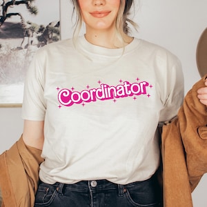 May include: A white t-shirt with a pink graphic that says "Coordinator" with stars around the text.