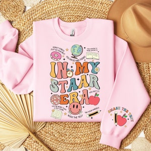 May include: Pink sweatshirt with a graphic design that says "In My Staar Era" with colorful illustrations of a donut, a globe, a book, a clock, a disco ball, a smiley face, an apple, and a pencil. The text "Smash the Test" is on the sleeve.
