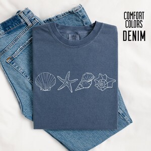 Può includere: Una maglietta blu denim con un design di conchiglie bianche, tra cui una conchiglia, una stella marina e due conchiglie. La maglietta è piegata sopra un paio di jeans blu. Il testo "COMFORT COLORS DENIM" è in nero.