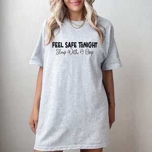 Puede incluir: Camiseta gris de gran tama&ntilde;o con el texto "Feel Safe Tonight Sleep With A Cop" impreso en negro.