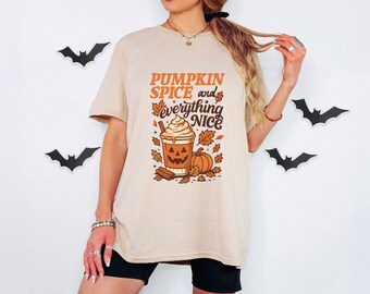 May include: Beige t-shirt with the text "PUMPKIN SPICE and everything NICE" in orange and white. The graphic includes a pumpkin spice latte, pumpkins, and fall leaves. Black shorts are worn with the shirt. Black bat decorations are on the white wall.