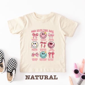 May include: A beige t-shirt with a graphic print of smiley faces and the text "GOD SAYS YOU ARE" with bible verses. The graphic includes the words "Beautiful, Victorious, Chosen, Redeemed, Strong, Amazing, Precious, Enough, Free".