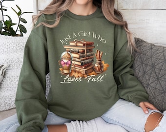 May include: Olive green sweatshirt with the text "Just A Girl Who Loves Fall". The graphic features a stack of books, a coffee drink with a butterfly, and cinnamon sticks. The design is in shades of brown, orange, and pink.