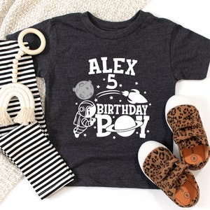 May include: A dark grey t-shirt with a white astronaut, planets, and stars design. The text "Alex 5 Birthday Boy" is printed on the shirt.