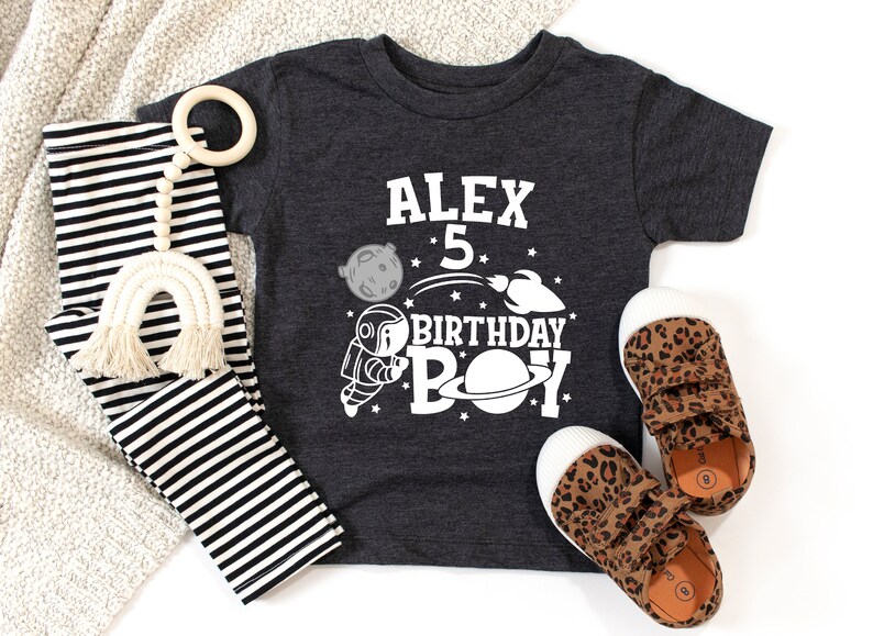 May include: A dark grey t-shirt with a white astronaut, planets, and stars design. The text "Alex 5 Birthday Boy" is printed on the shirt.
