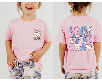 May include: Pink t-shirt with a baseball graphic and the text "In My Baseball Sister Era" on the back. The front of the shirt has a baseball graphic with a bow and the name "Sarah" printed below.