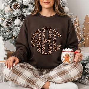 May include: A brown sweatshirt with a leopard print monogram design. The sweatshirt is paired with plaid pants. A festive mug with a gingerbread man is held in the hand. The background includes a decorated Christmas tree and wooden tree decorations.