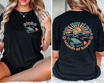 May include: Black t-shirt with "aloha" and a turtle graphic on the front. The back has a circular design with "aloha state of mind islands of hawaii", a sun, waves, and a turtle. The design evokes a Hawaiian theme.