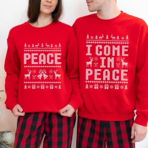 May include: Matching red Christmas sweaters with white text and festive patterns. One sweater says "PEACE", the other says "I COME IN PEACE". Both are paired with red and black plaid pajama pants, perfect for holiday gatherings.