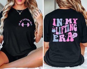 May include: Black t-shirt with the text "IN MY LIFTING ERA" in pink and blue letters with a dumbbell graphic on the front and a kettlebell graphic on the back.