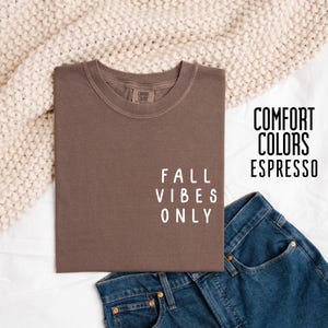 Può includere: Una maglietta marrone con le parole "FALL VIBES ONLY" stampate in bianco. La maglietta è abbinata a jeans blu. È visibile anche il testo "COMFORT COLORS ESPRESSO".