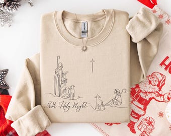 Op de afbeelding: Beige crewneck sweatshirt met een minimalistische lijntekening van een kerststal en de tekst "Oh Holy Night". Het ontwerp bevat figuren, een kruis en schapen. Een ketting met een zonnehanger is zichtbaar.