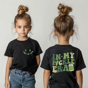 May include: A black t-shirt with a green and white graphic that says "In My Pickle Era" on the back and a green and white smiley face with the text "Pickle Era" on the front.