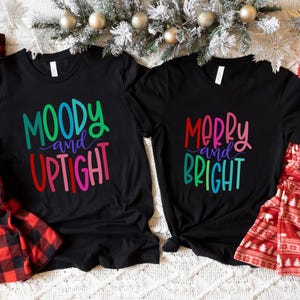 May include: Two black t-shirts with colorful text that reads "Moody and Uptight" and "Merry and Bright". Red and black plaid pajama pants and red pajamas with a white bear pattern are also visible. A white mug is also present.