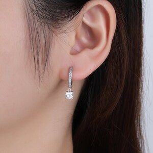 May include: Silver hoop earrings with a single clear stone dangling from the bottom. The hoop is lined with small clear stones.
