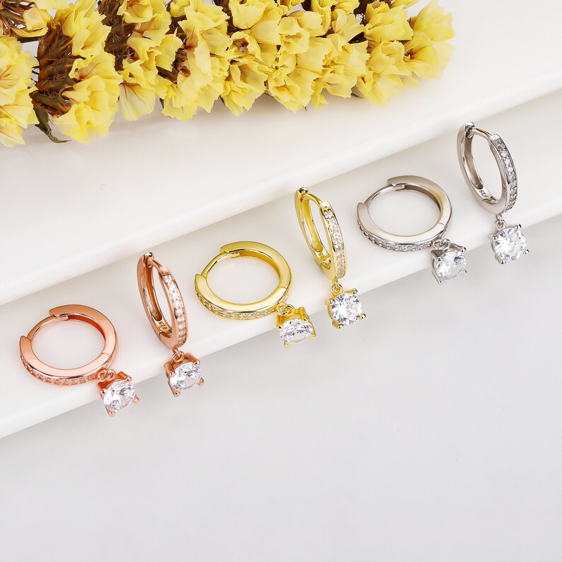 May include: Five pairs of hoop earrings with a dangling crystal. The hoops are available in gold, silver, and rose gold.