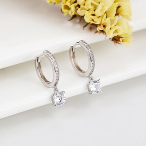 May include: A pair of silver hoop earrings with a dangling round clear stone. The hoops are accented with small clear stones.