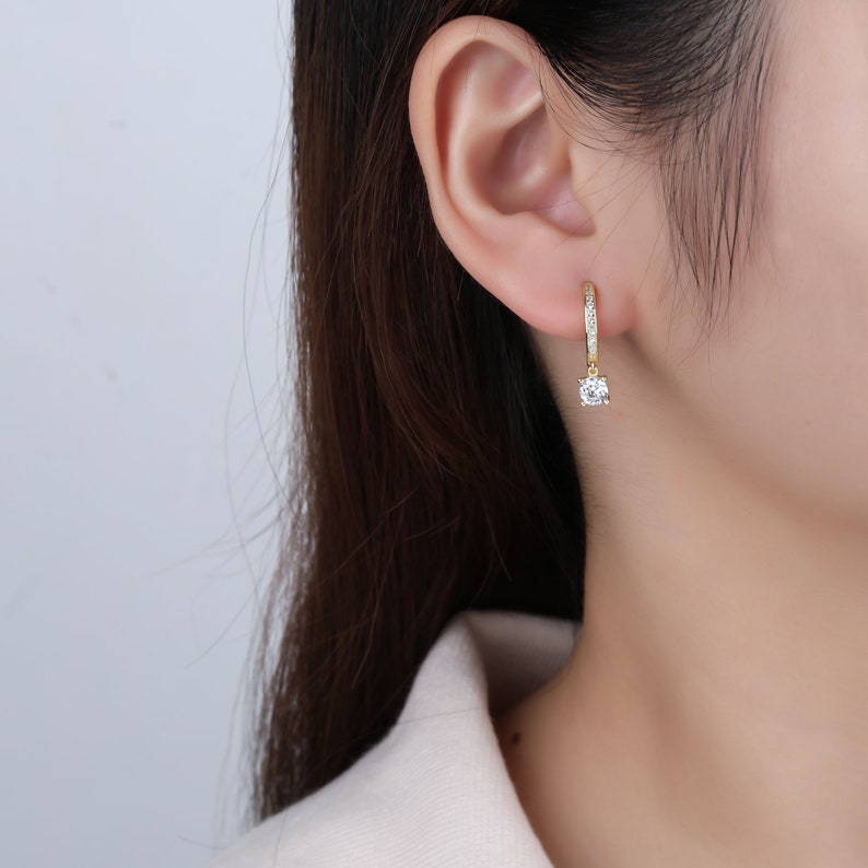 May include: Gold hoop earrings with a single clear stone dangling from the bottom. The earrings are set with small clear stones around the hoop.
